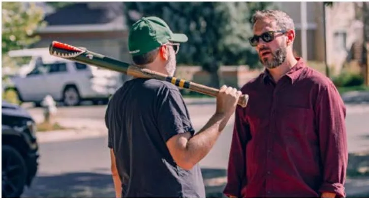 Two men talk on a residential street; one holds a baseball bat on his shoulder.